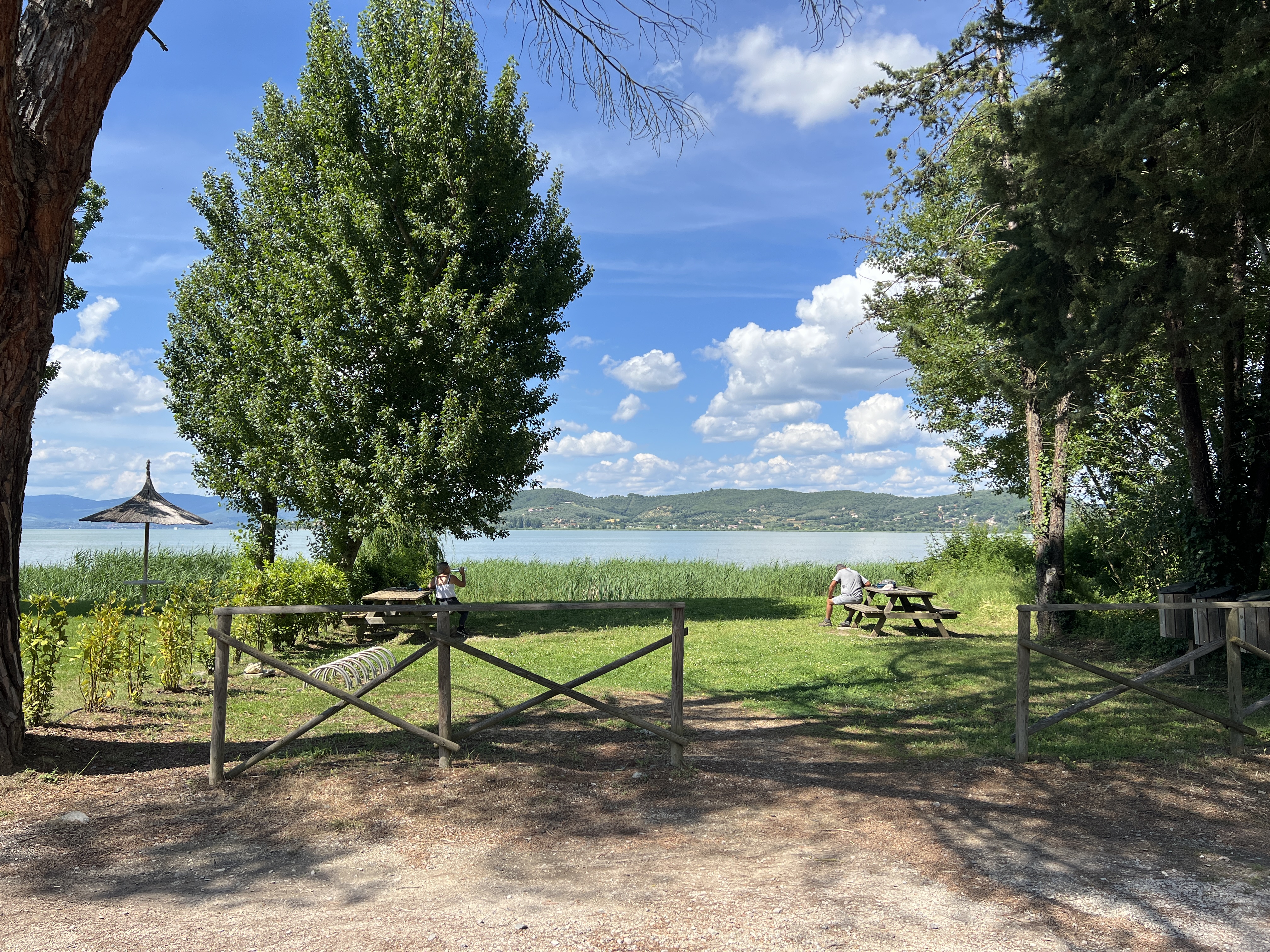 Green area along lake, with picnic table, benches, bike racks and views of Lake Trasimeno and hills.